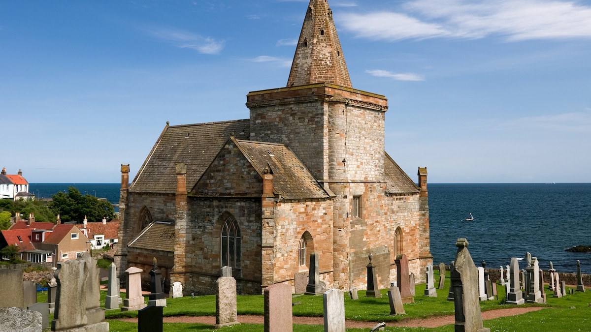 A historic church is situated near the waterfront, surrounded by a cemetery with various gravestones on grassy grounds. The stone building features a distinct tower and large arched windows. The coastline and sea are visible in the background, under a clear blue sky.