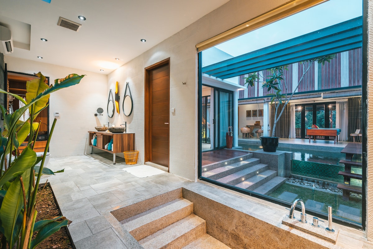 A welcoming entrance area features contemporary decor, including decorative mirrors and a stylish console table. The space is illuminated by natural light streaming through large windows, revealing a glimpse of the private outdoor area with landscaped greenery and outdoor seating.