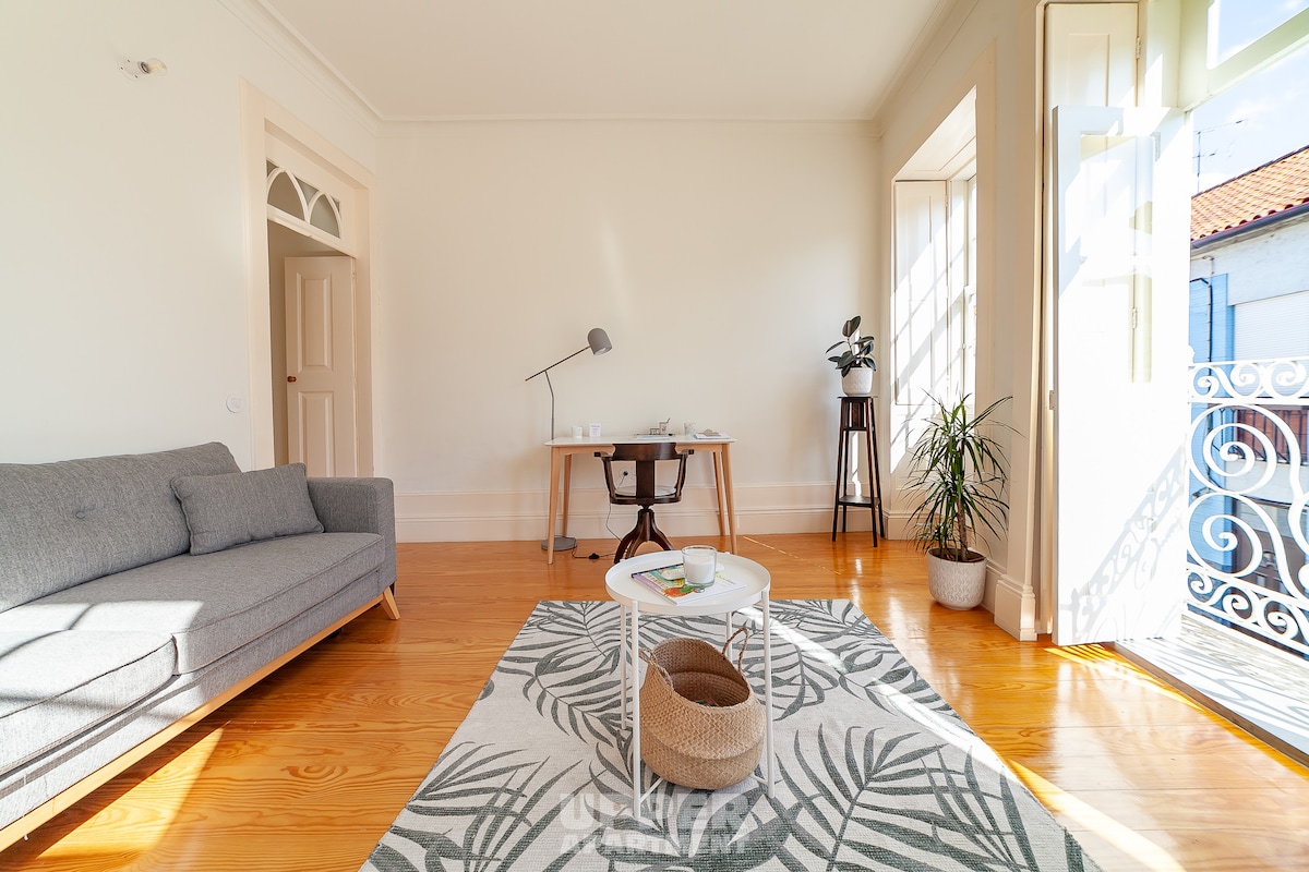 A spacious living area features a light grey sofa, a circular coffee table, and a patterned rug. Sunlight fills the room through large windows, illuminating a small desk with a chair and a potted plant. A French door opens to the outside balcony.