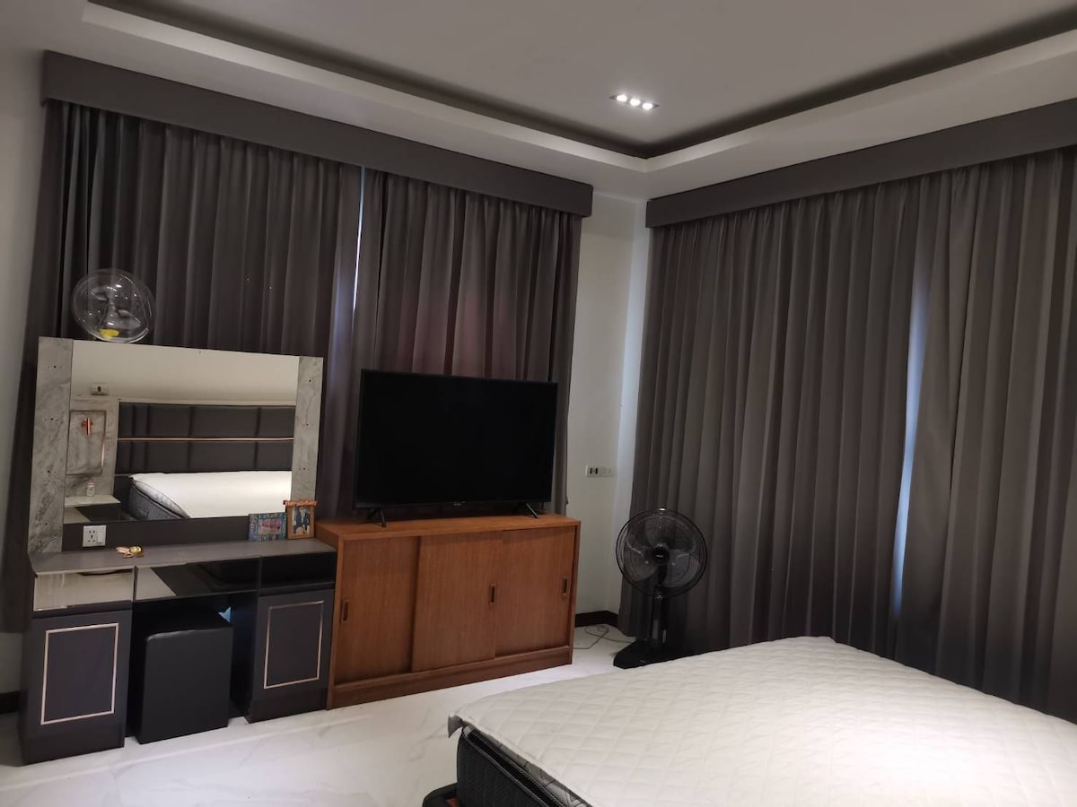 The room features light-colored flooring and modern furnishings. A spacious bed is partially visible, complemented by dark curtains that frame the windows. A television stands on a sleek console alongside a fan. A mirrored dresser adds depth to the space.