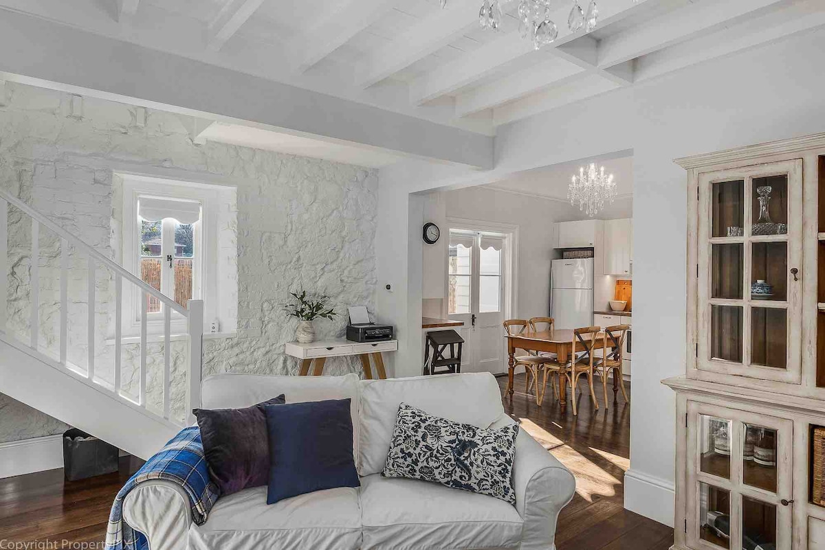 A light-filled interior features a comfortable white sofa adorned with dark blue and patterned cushions. Exposed wooden beams highlight the living area, while a dining table and chairs are visible in the background. Textured stone walls add character to the space.