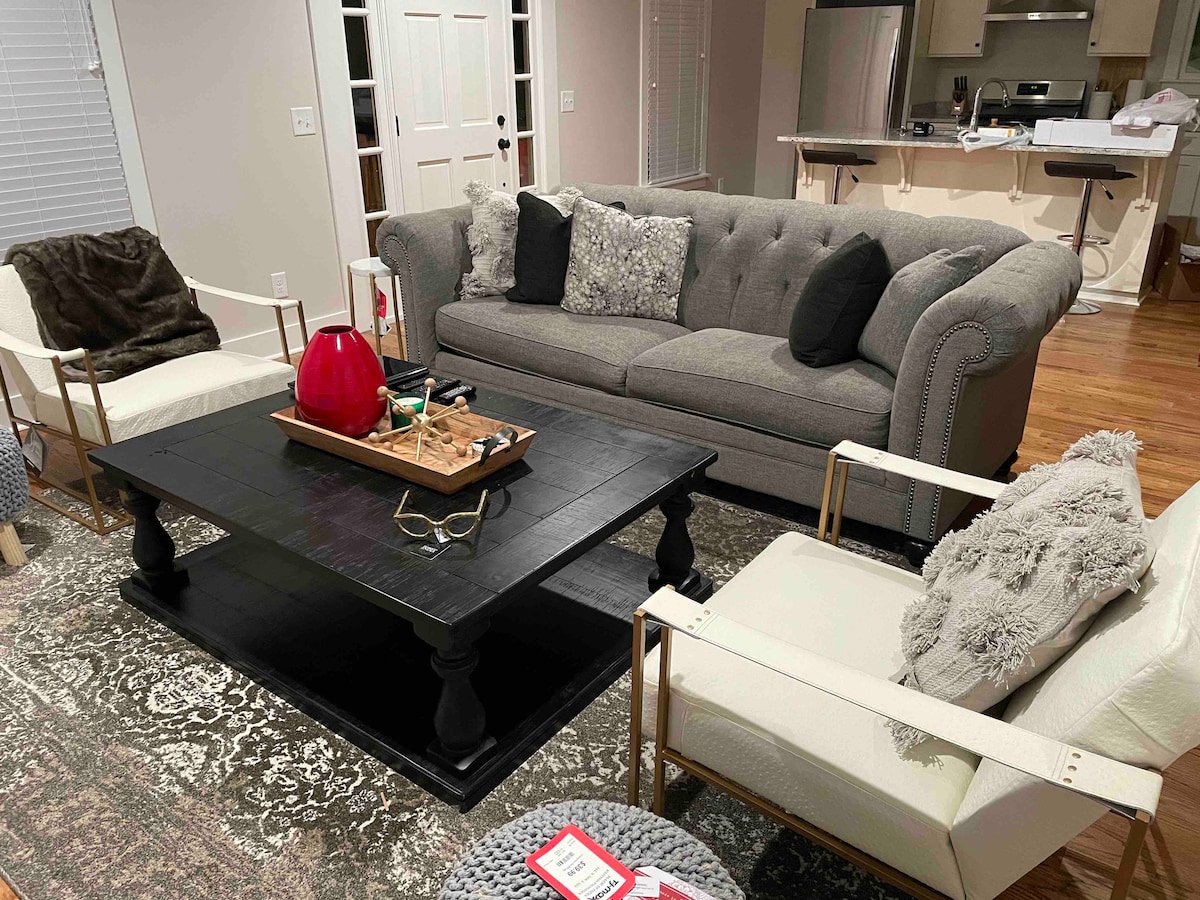 A comfortable living area is presented with a plush gray sofa featuring decorative cushions. A dark wooden coffee table occupies the center, adorned with decorative items. Two modern white chairs provide additional seating, and a patterned area rug adds texture to the wooden floor.