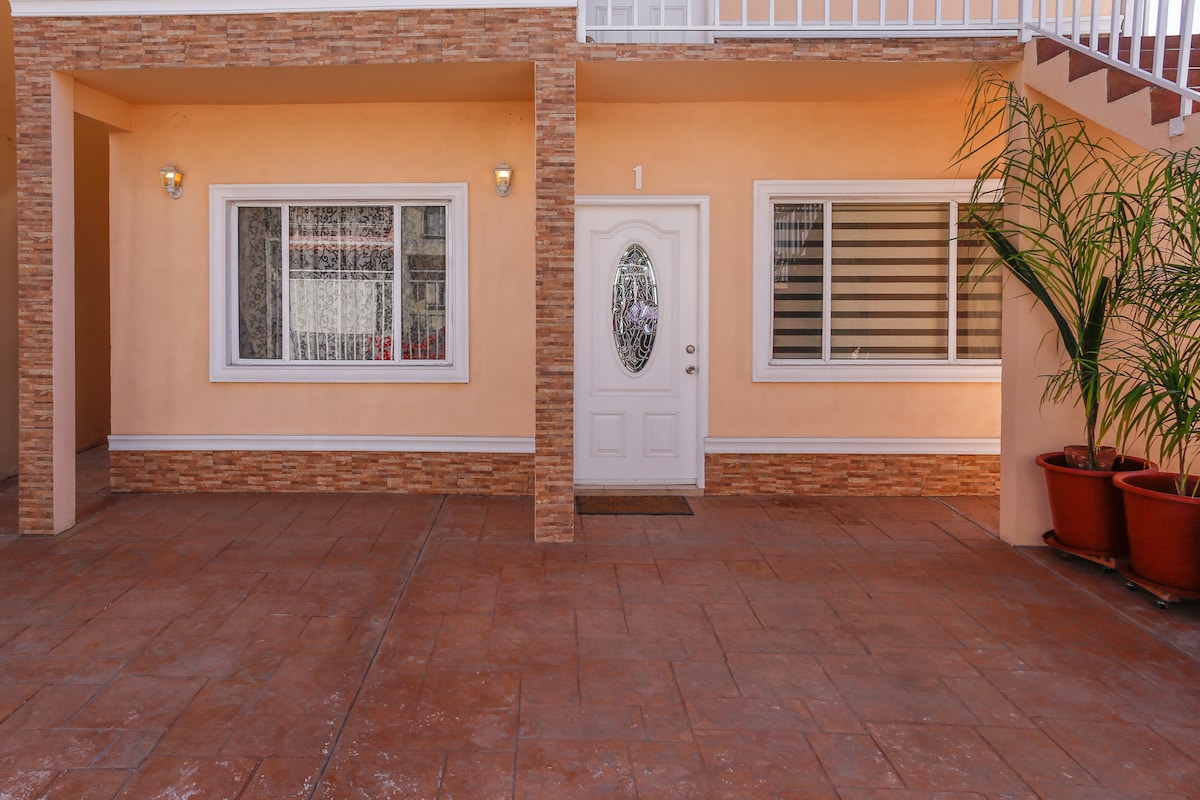The exterior entrance features a white door framed by decorative stone accents. A large window beside the door allows light to enter, with sheer curtains softly diffusing it. Potted plants in terracotta planters are positioned on either side of the entrance, contributing to a welcoming appearance.