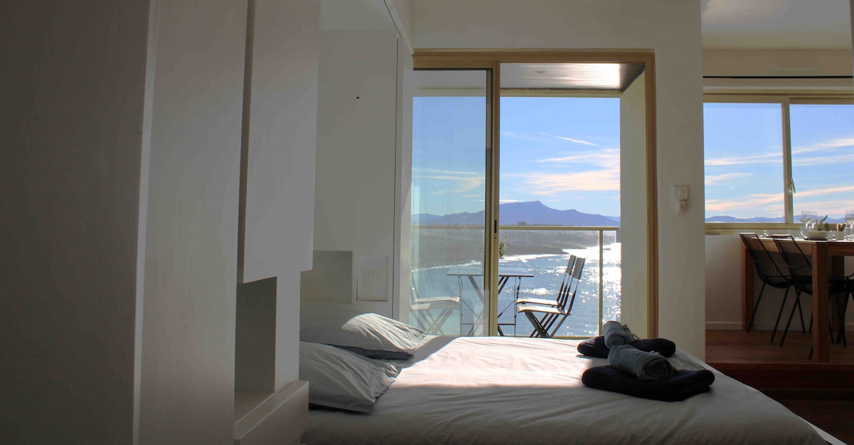 A cozy sleeping area features a spacious bed with blue bedding, positioned near sliding glass doors. A private balcony offers a scenic view of the coastline and mountains. A dining area can be seen in the background, accommodating a table and chairs.