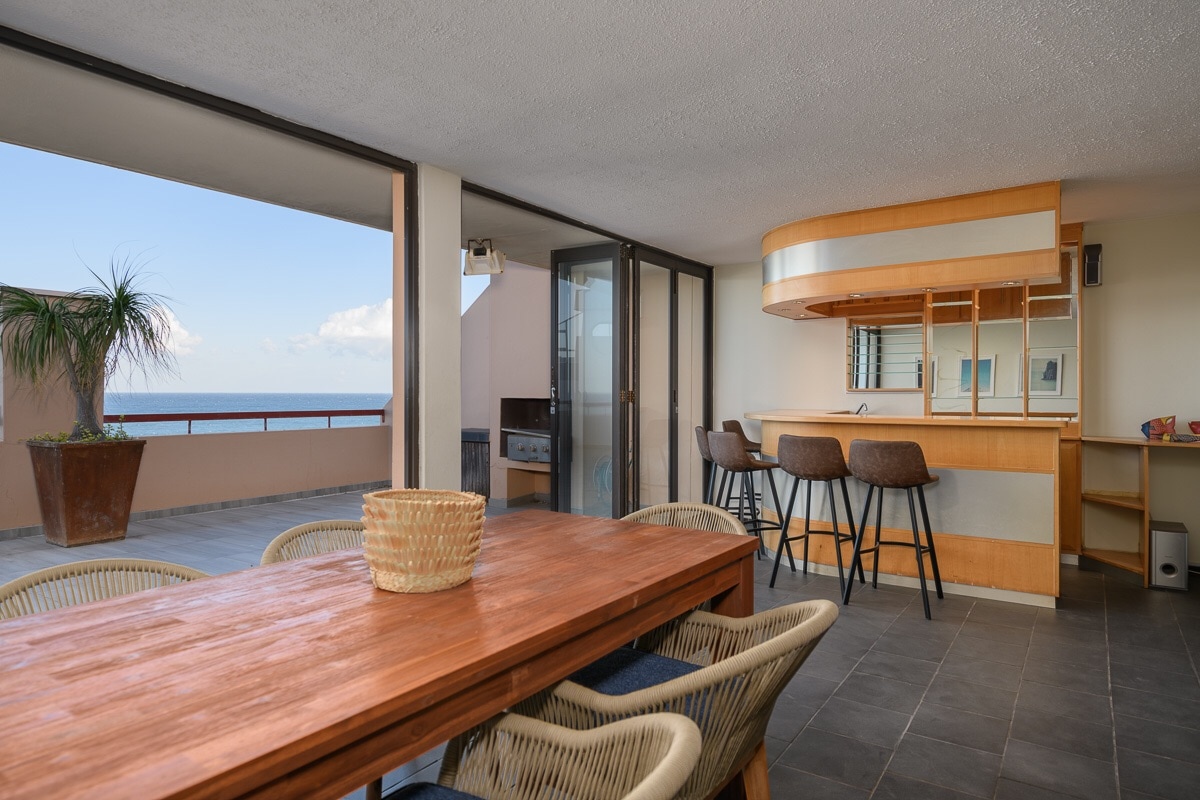 An open dining area features a wooden table surrounded by woven chairs. A sleek bar counter with seating is positioned nearby, offering views of the Indian Ocean through large glass doors. Natural light floods the space, highlighting the neutral tones of the decor.