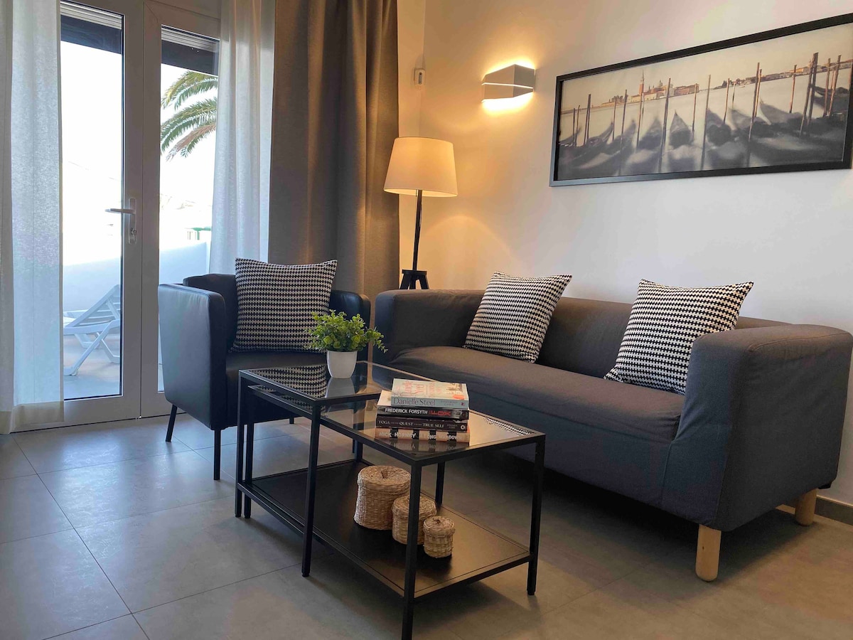 A comfortable seating area is visible, featuring a grey sofa with patterned cushions and a sleek glass coffee table. A side lamp provides gentle lighting. Light curtains frame the glass doors leading to a balcony, with a glimpse of outdoor furniture and palm trees in the background.
