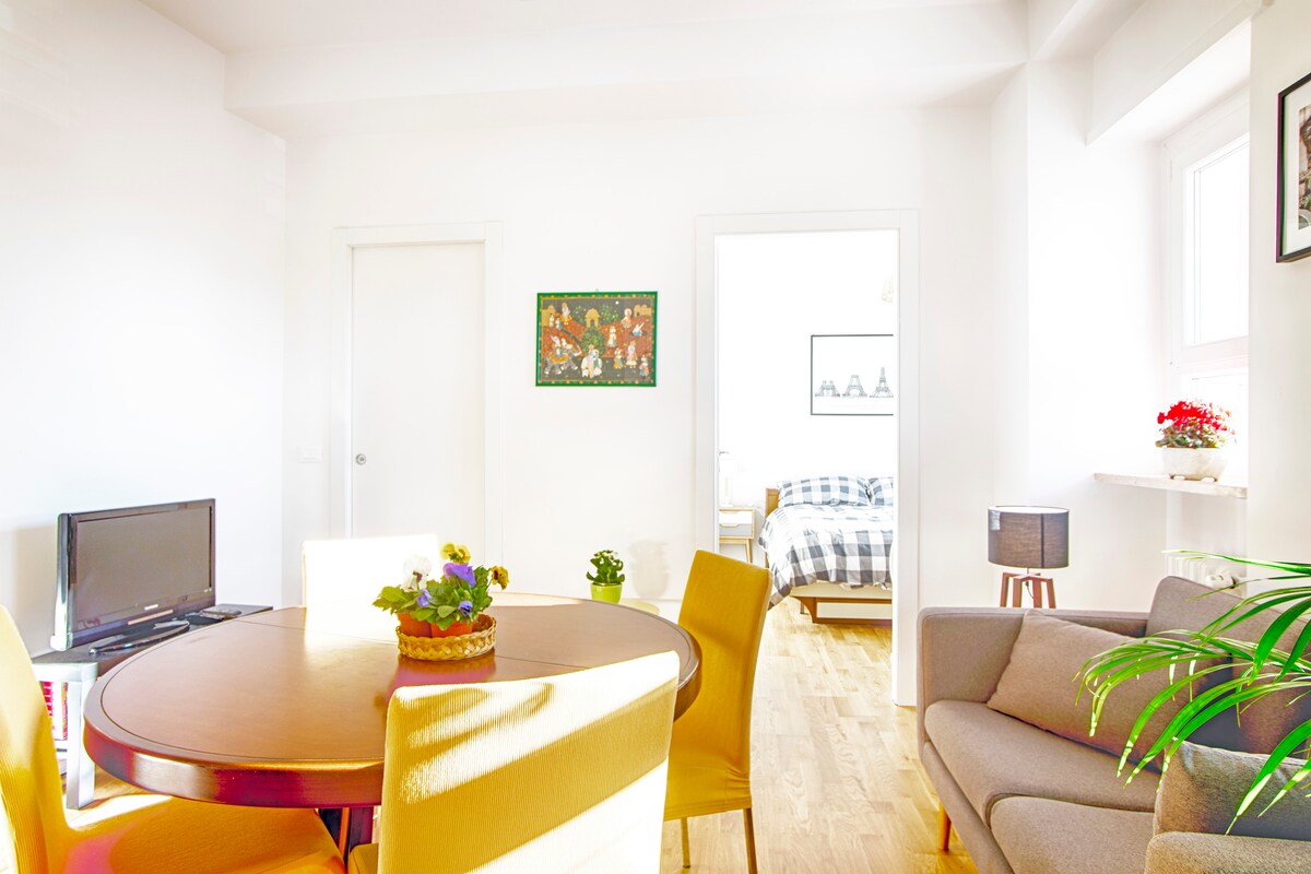 A bright living space features a round wooden table surrounded by yellow chairs. A television is situated on a stand to the left, while a cozy gray sofa provides seating. Natural light fills the room, highlighting vibrant wall art and a small potted plant.