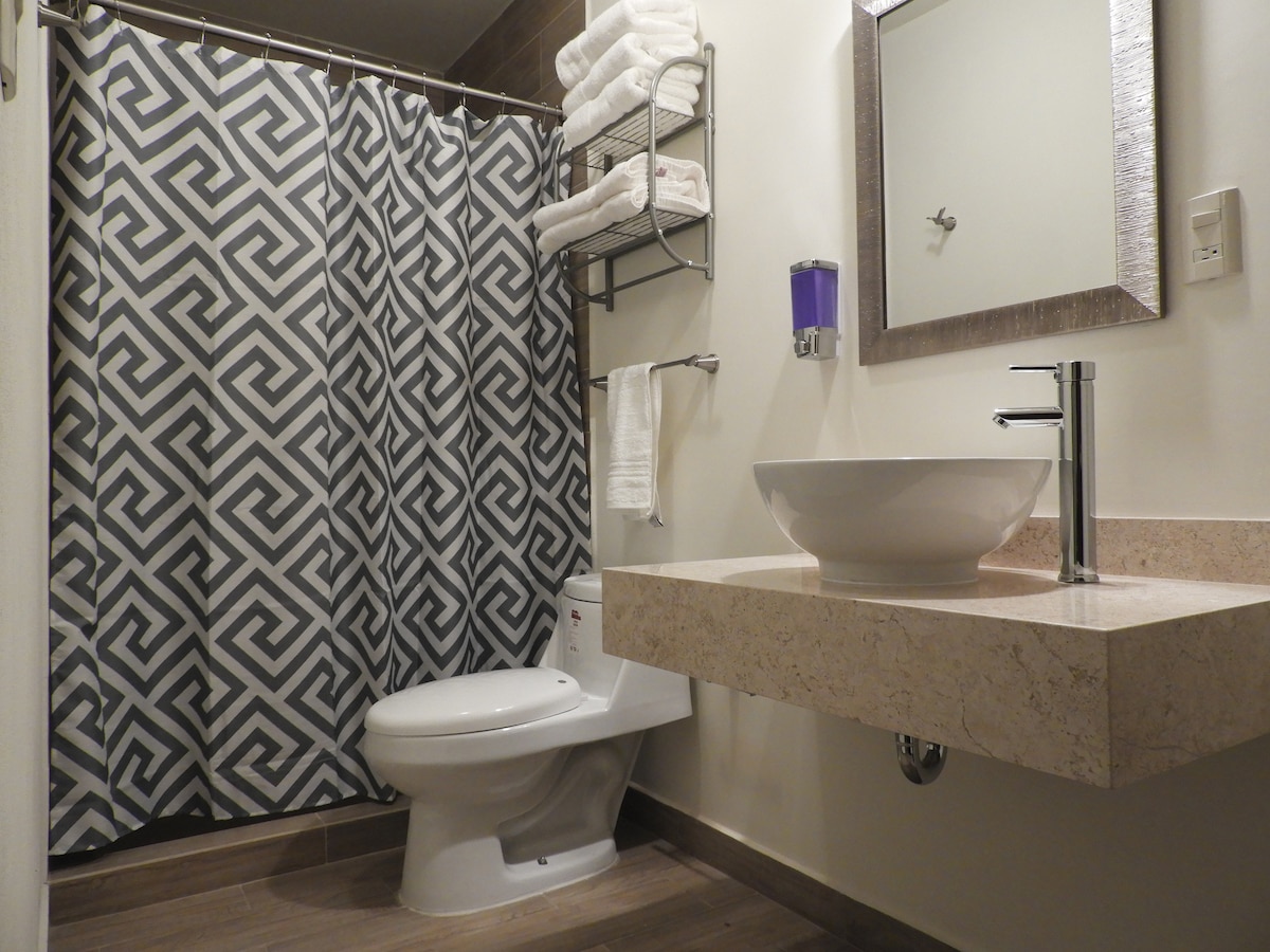 A modern bathroom features a stylish gray and white patterned shower curtain, a wall-mounted shelf with neatly arranged towels, and a sleek sink with a minimalist faucet. Natural wood tones are present in the flooring, enhancing the overall clean and contemporary aesthetic.