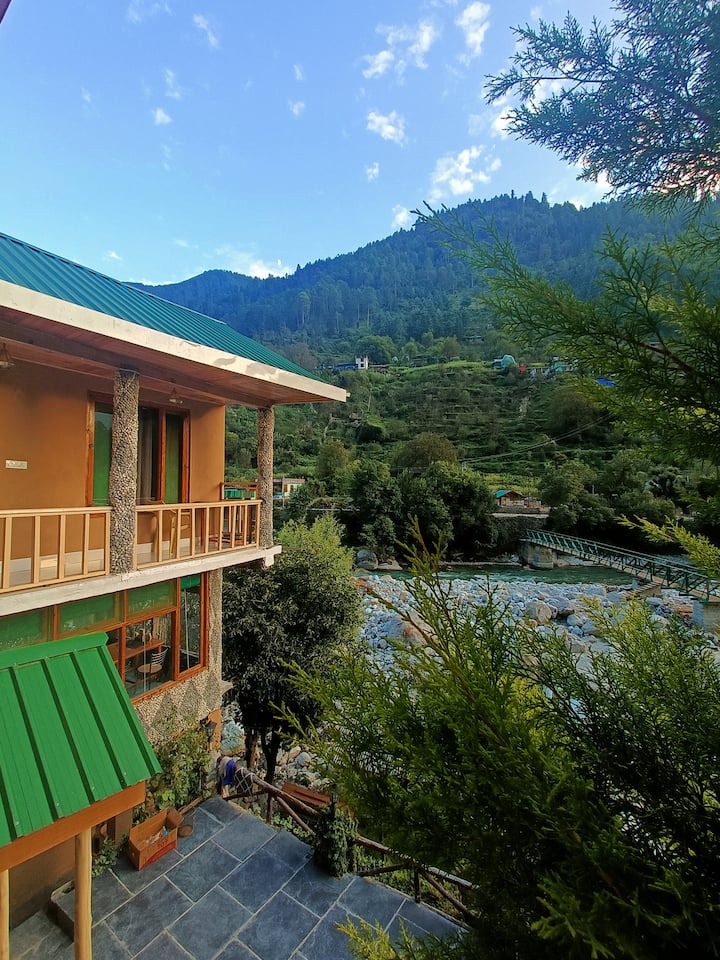 The house from across the river — green bridge, forest slopes, and the Tirthan carving its path through the valley