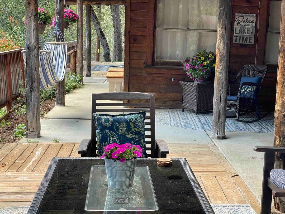 A covered front deck is presented, featuring a wooden table set with a potted plant. Comfortable seating is arranged around the table, with a hammock visible nearby. Colorful flowers are displayed in decorative planters, enhancing the outdoor space.