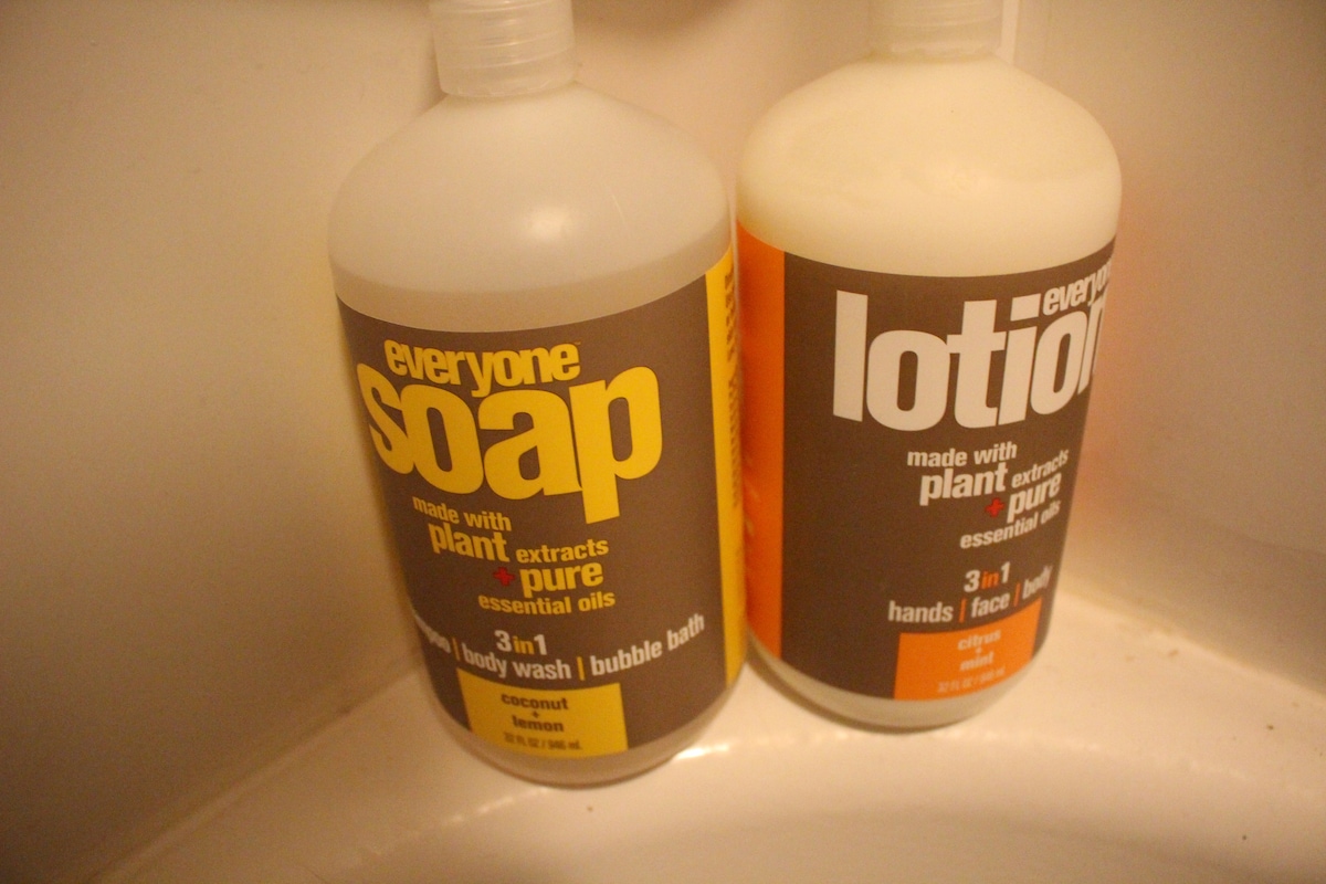 Two bottles of personal care products are positioned on a countertop. One bottle contains body wash, labeled with coconut and lemon scents. The second bottle features lotion, also labeled with instructions for hands and face use, boasting a cream-colored design with orange accents.