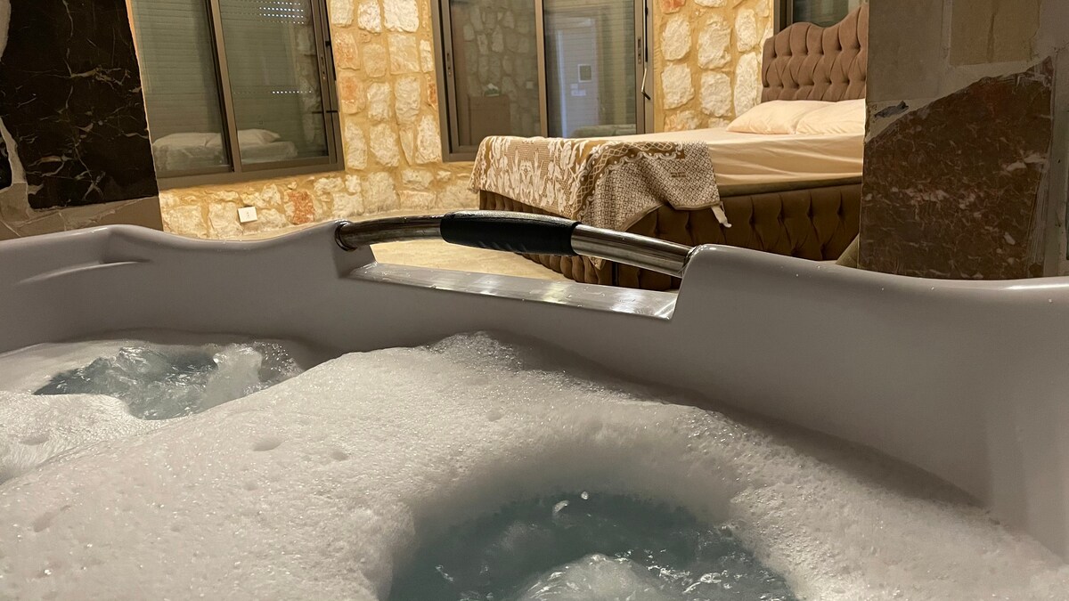 A bubbly hot tub is shown in the foreground, with jets creating a soothing effect. In the background, a spacious bed with a tufted headboard is partially visible, contributing to a relaxing retreat ambiance. Stone walls create a warm atmosphere.