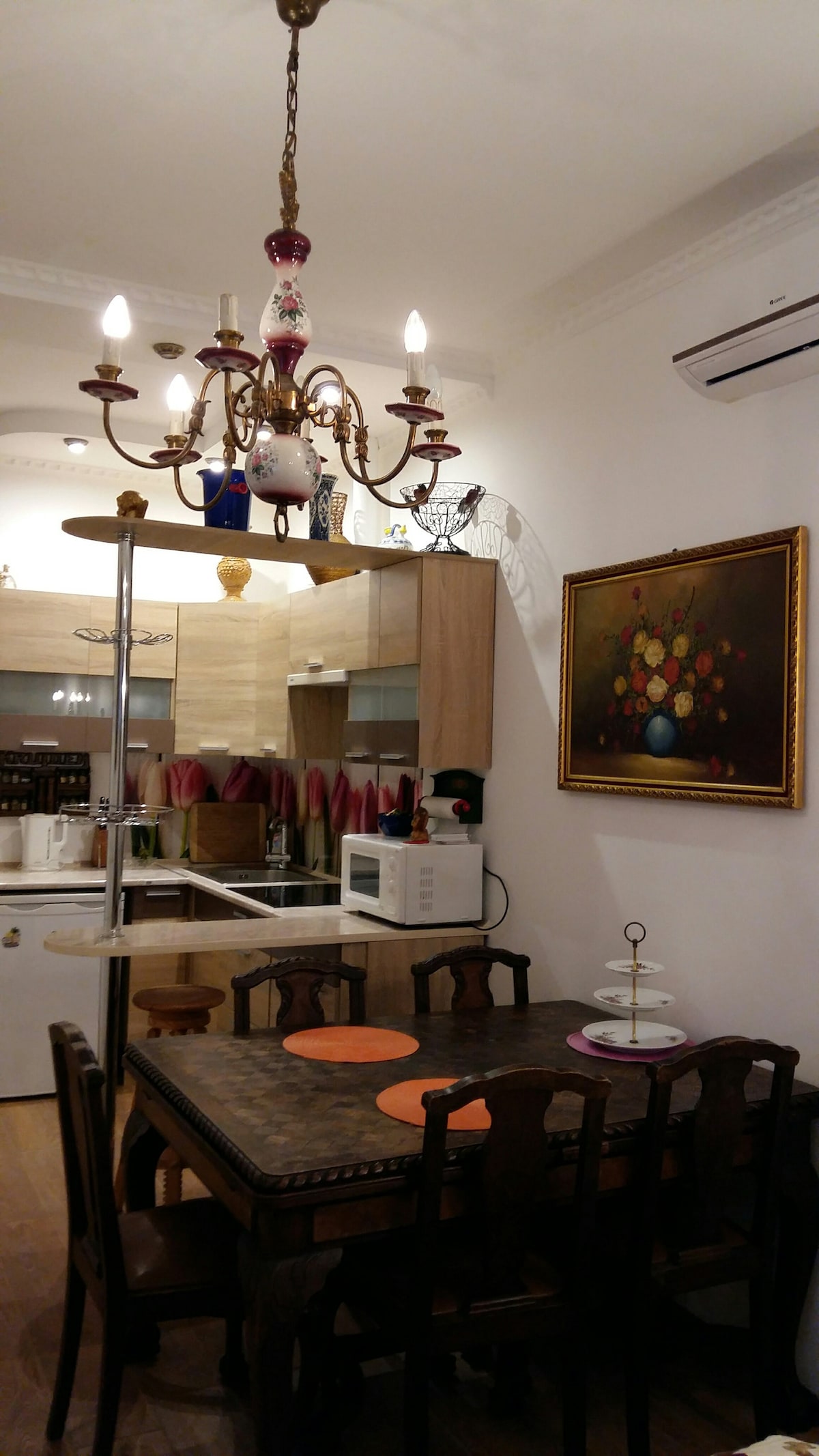 A dining area features a wooden table surrounded by high-backed chairs. A chandelier hangs above, providing ambient light. A colorful painting with floral motifs is displayed on the wall, while a microwave is positioned on a countertop, enhancing the functionality of the space.
