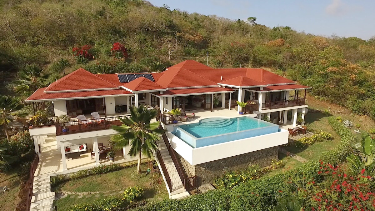 An aerial view of a spacious residence with a red-tiled roof is presented, showcasing a large infinity pool at the edge of the property. Lush gardens surround the home, with vibrant tropical plants and trees visible throughout the landscape.