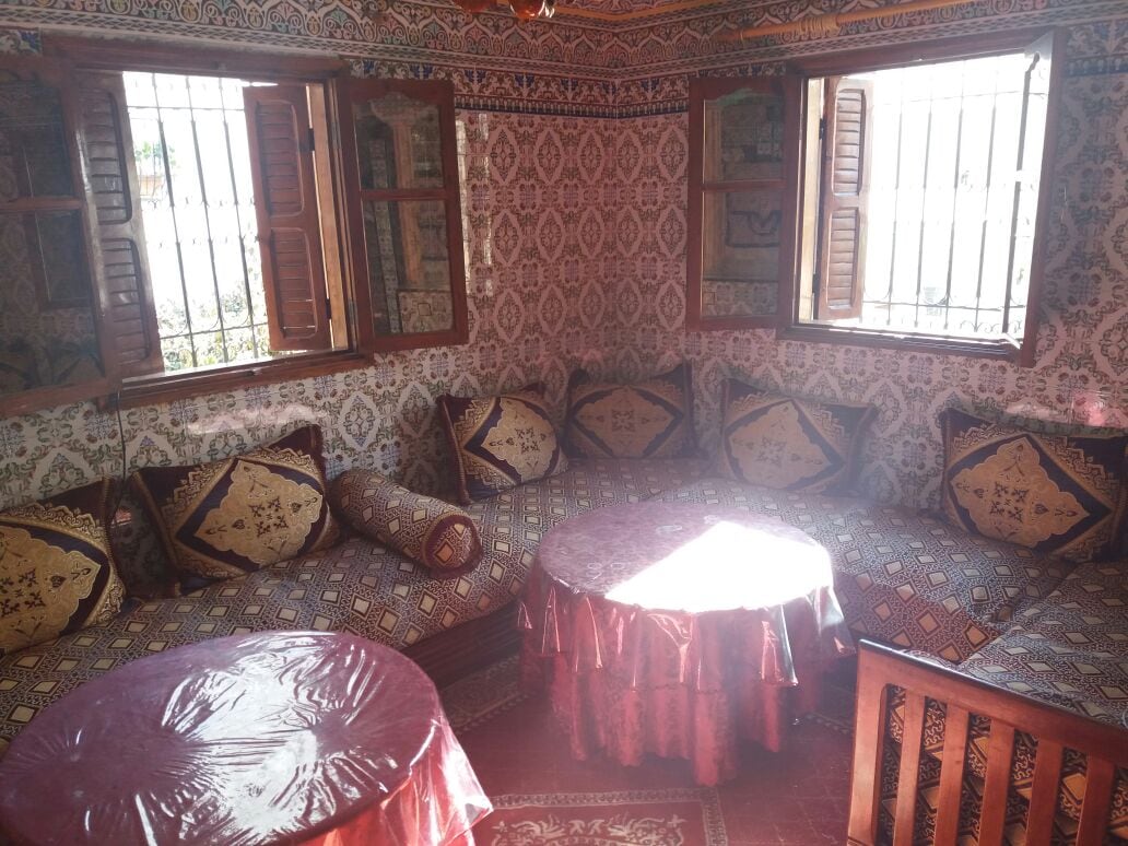 A cozy seating area is adorned with intricate patterned textiles and decorative pillows. Two round tables are positioned in front of a spacious low seating arrangement that surrounds the room. Natural light filters through the window bars, illuminating the vibrant decor.