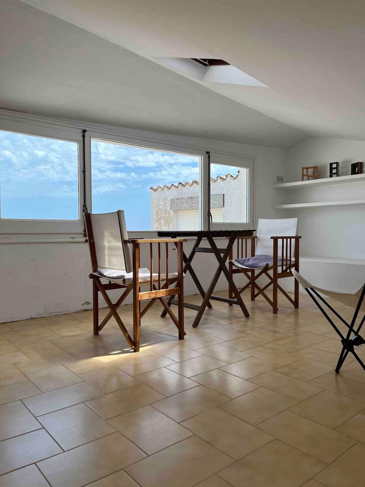 A bright attic room is presented, featuring a wooden table surrounded by four chairs. Large windows provide natural light and views of the outdoors. Light-colored tiles adorn the floor, and a shelf on the wall showcases decorative items.
