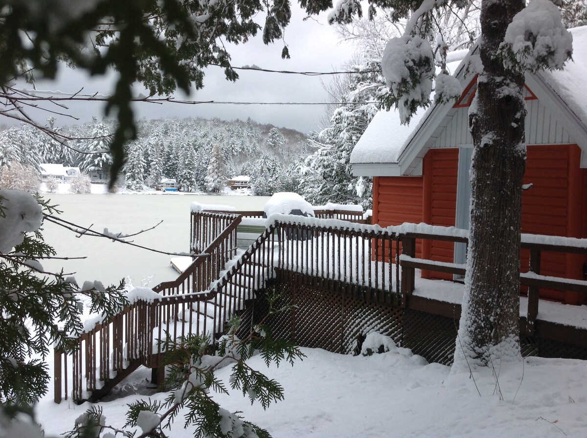 A cozy chalet is framed by snow-covered trees, showcasing a wooden deck that leads toward the lake. Fresh snow blankets the surrounding landscape, and a tranquil view of the frozen lake is visible in the background, highlighting the serenity of winter.