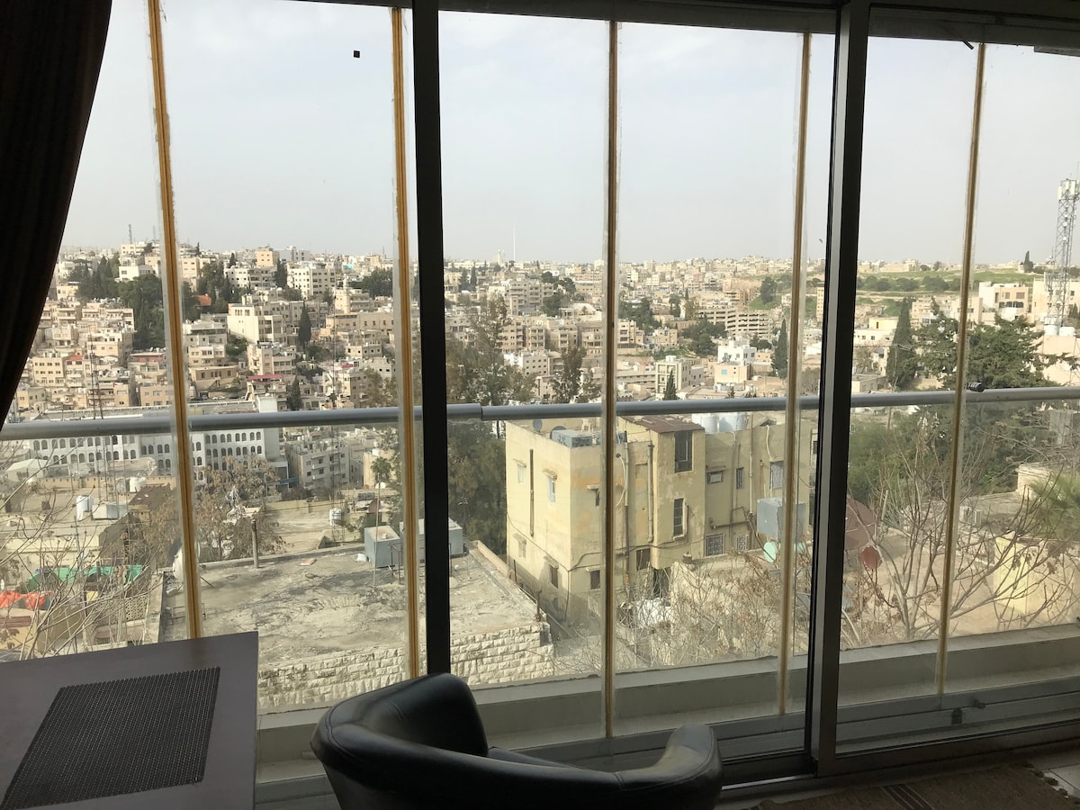 A panoramic view of the Amman skyline is framed by large glass windows, allowing natural light to fill the space. The bustling cityscape stretches out, showcasing a mix of traditional and modern architecture, with hills visible in the distance.