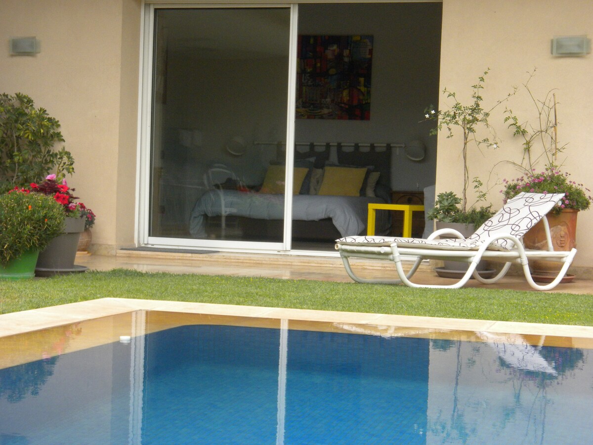 A modern bedroom is visible through large glass doors, featuring a spacious bed with light bedding and colorful accent pillows. A bright yellow side table adds a pop of color. In the foreground, a sleek lounge chair rests beside a clear pool, surrounded by lush greenery and blooming flowers.