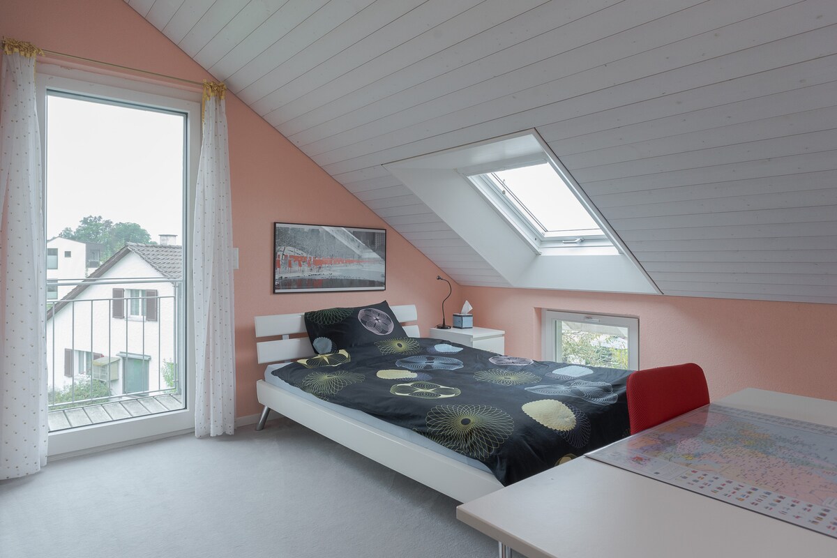 A bright guest room features a spacious bed adorned with a patterned duvet. Large windows bring in natural light, and a small table with chairs is located nearby. The walls are painted in a soft pink hue, enhancing the room's inviting ambiance.