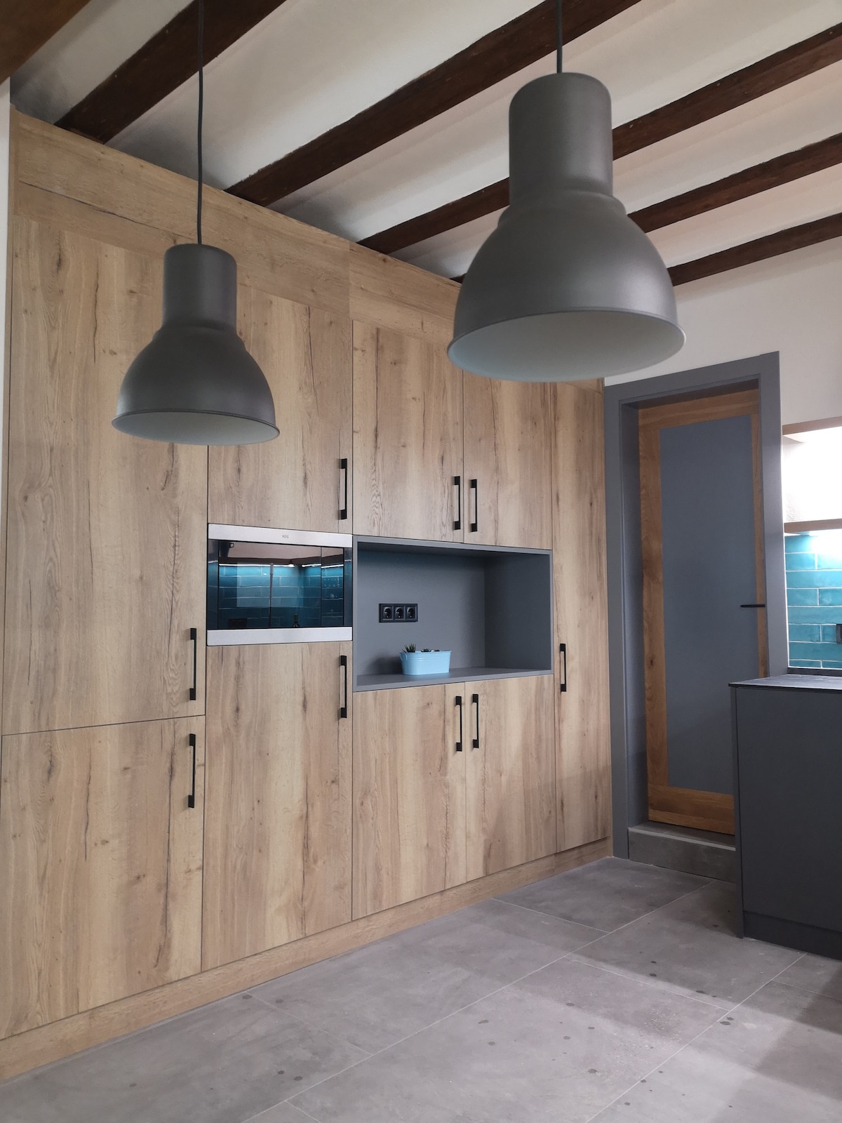 A modern kitchen area is showcased with wooden cabinetry featuring sleek handles. Two gray pendant lights hang from the ceiling above, illuminating the space. A gray countertop is visible, complemented by a minimalistic design and large windows providing natural light.
