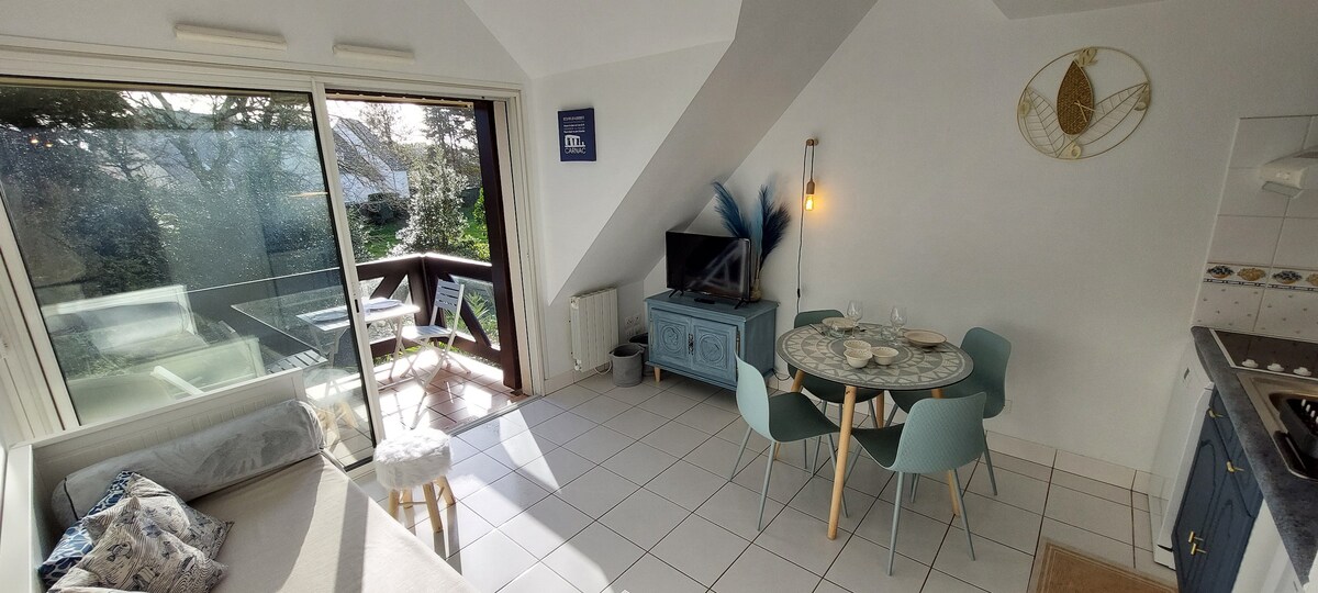 A well-lit living area showcases a dining table with light blue chairs and a television on a side cabinet. Large glass doors open to a terrace, while a comfortable sofa and decorative elements add to the relaxed ambiance.