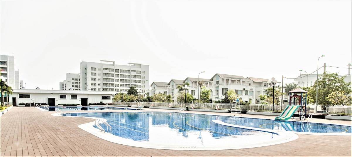 A spacious swimming pool is visible, featuring clear blue water and surrounded by wooden decking. The area includes a slide and lounge chairs, with well-maintained garden spaces and several multi-story buildings in the background, indicating a serene recreational environment.