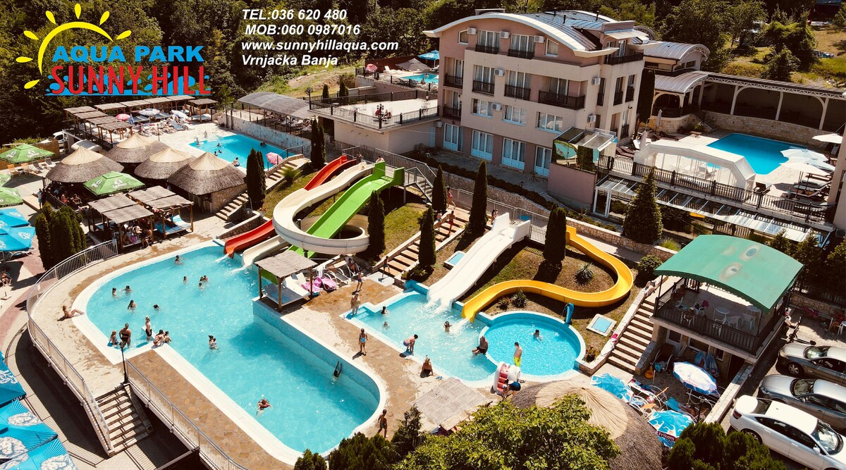 An aerial view captures the vibrant water park at Sunny Hill Spa Resort, featuring several swimming pools and colorful water slides. Guests can be seen enjoying the various water attractions, surrounded by lush greenery and lounge chairs positioned around the pool areas.