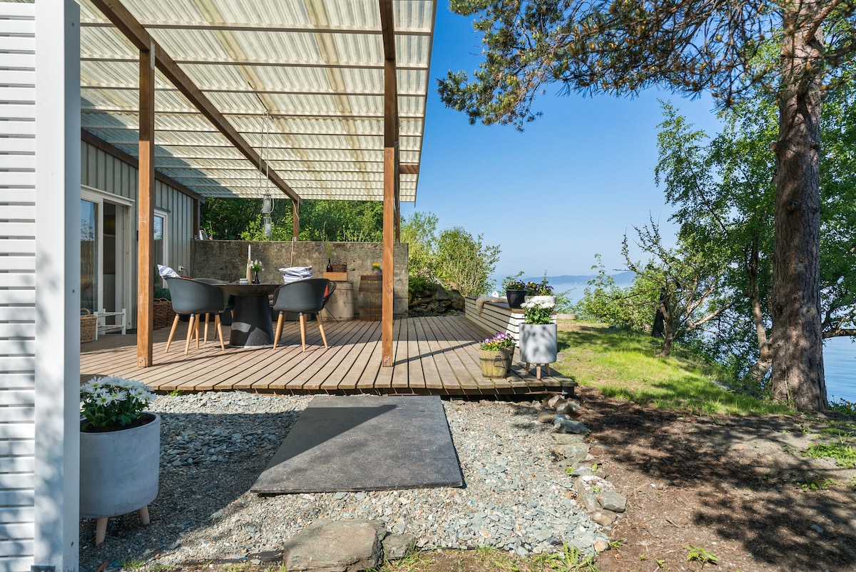 An outdoor deck features a shaded dining area with a table and chairs, surrounded by greenery and flowers. Scenic views of the fjord are visible from the deck, while the space is complemented by natural stones and a planting pot. The tranquil setting invites relaxation.