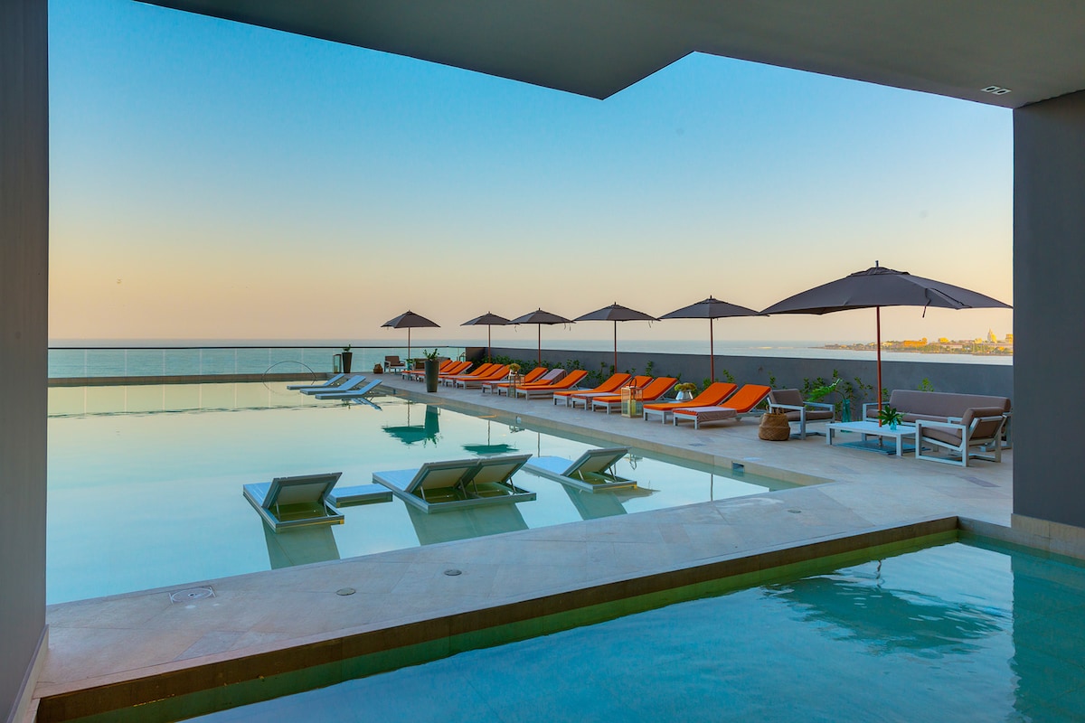 An outdoor pool area is visible, featuring several lounge chairs with umbrellas. The water reflects the colors of the sky, creating a serene atmosphere. Views of the ocean are seen in the distance, enhancing the relaxing nature of the space.