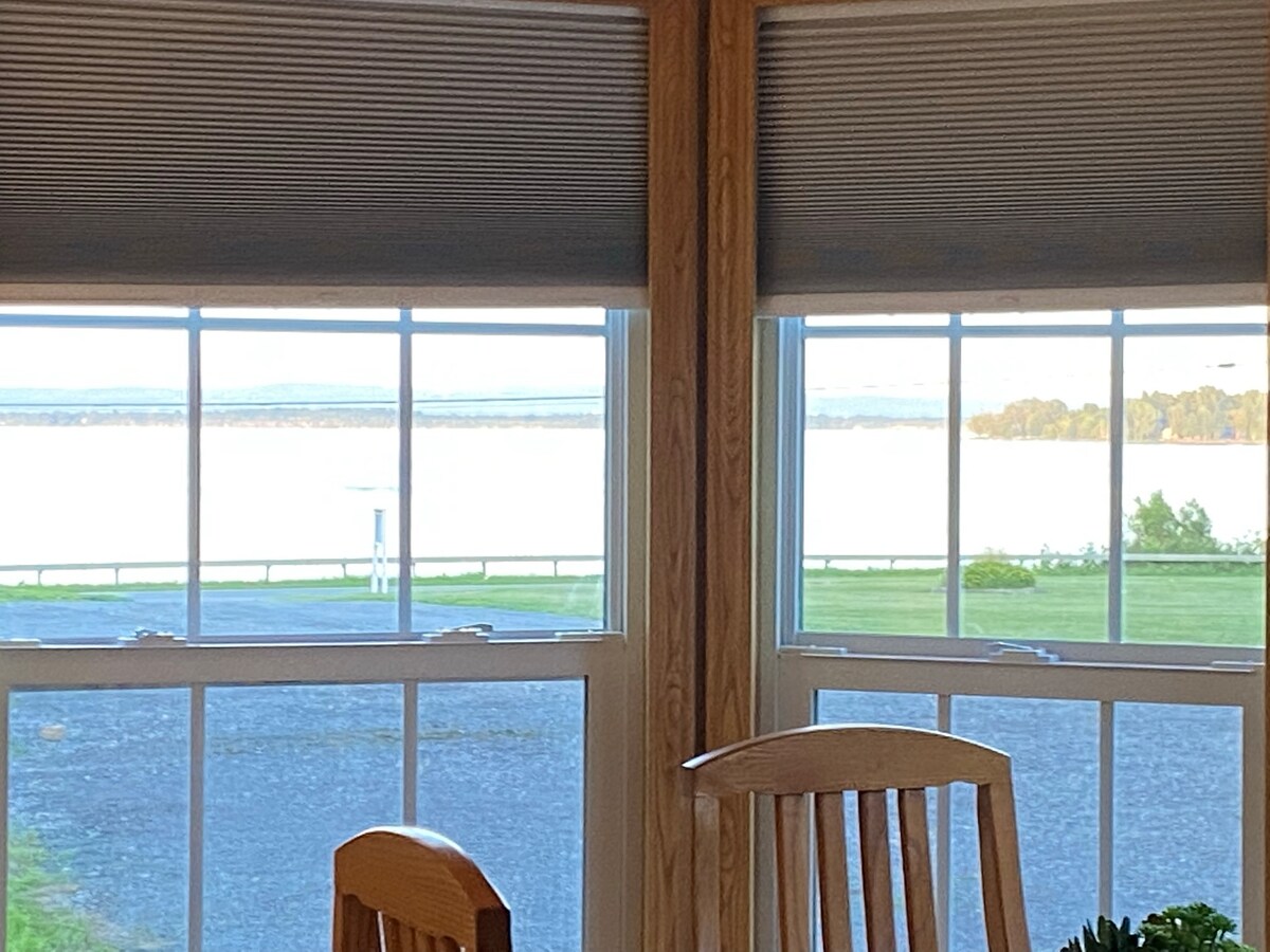 Large windows provide a view of the tranquil Lake Champlain, with daylight reflecting off the water. The blinds are partially raised, allowing natural light to fill the space, while a wooden dining table is positioned in front of the windows.