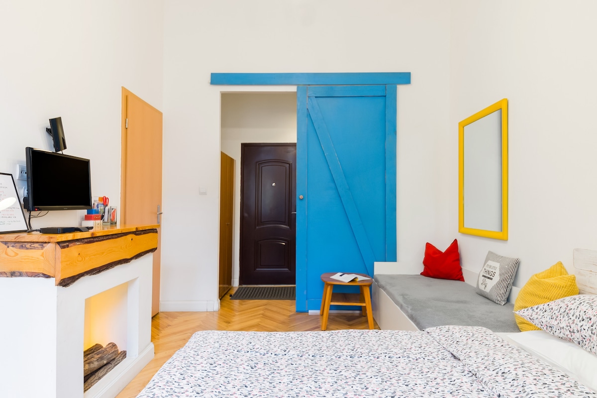 A bright, airy room features a cozy sleeping area with floral-patterned bedding. A grey sofa with colorful cushions complements the space. A blue sliding door adds a pop of color, while a wall-mounted TV and a wooden shelf are positioned near the entrance.