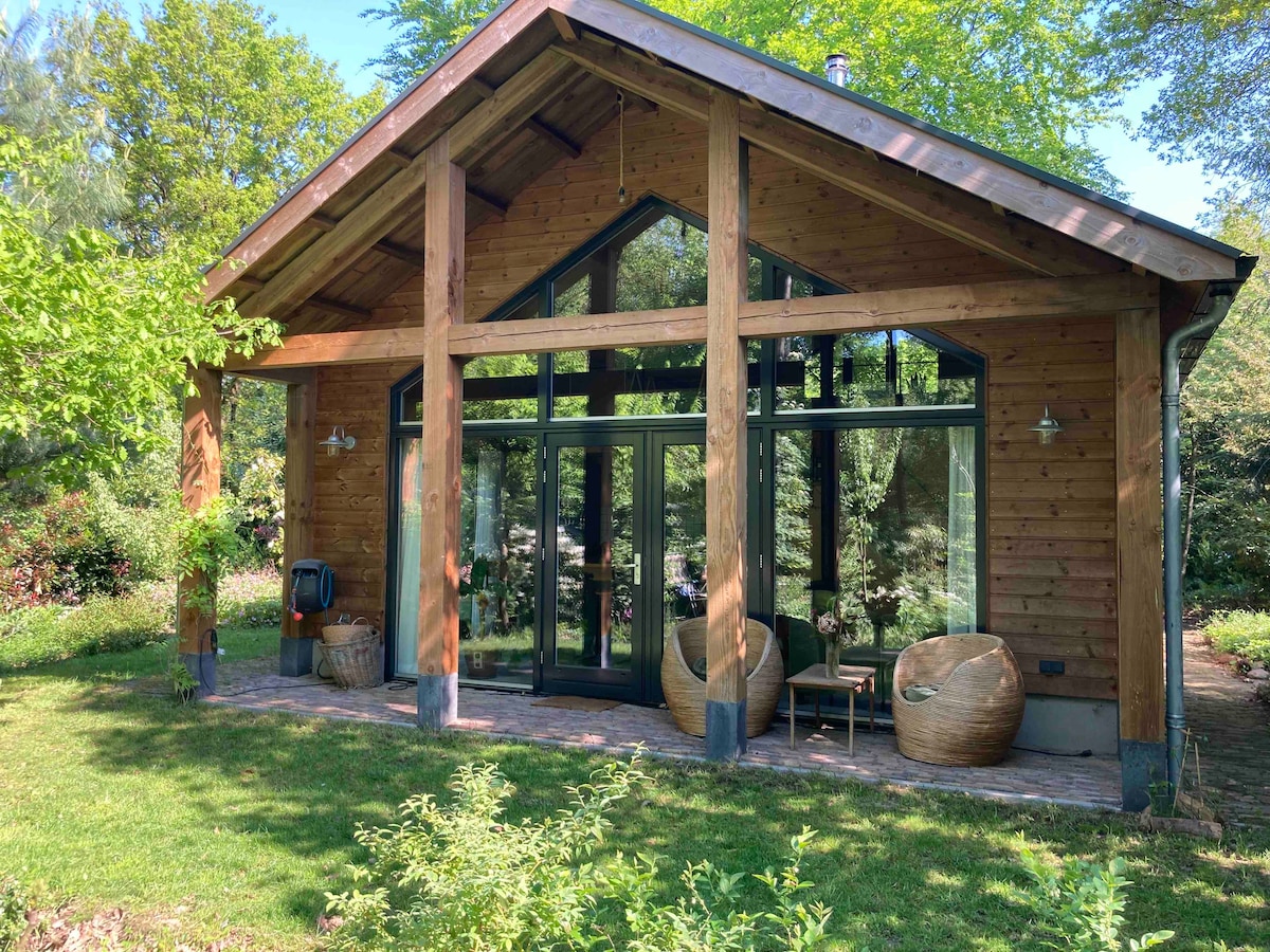 The wooden house features a large front porch with two woven chairs and a small table, set against a backdrop of lush greenery. Expansive glass doors invite natural light, showcasing a harmonious connection between indoor and outdoor spaces. Tall trees frame the setting, enhancing the serene atmosphere.