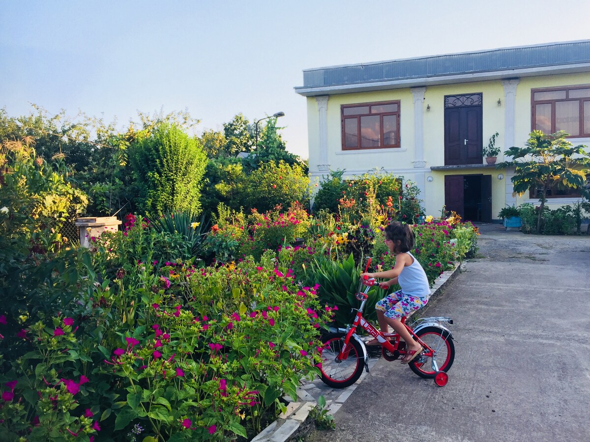 A child rides a red bicycle along a pathway lined with vibrant flowers in a garden. The home, with large windows and a wooden front door, is visible in the background, surrounded by lush greenery and colorful blossoms.