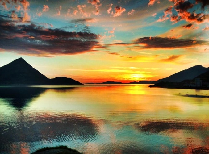 A breathtaking landscape captures a vibrant sunset over a tranquil body of water. The sky is painted with hues of orange, yellow, and purple, reflecting on the still surface below. Silhouetted mountains create a serene backdrop, enhancing the natural beauty of the scene.