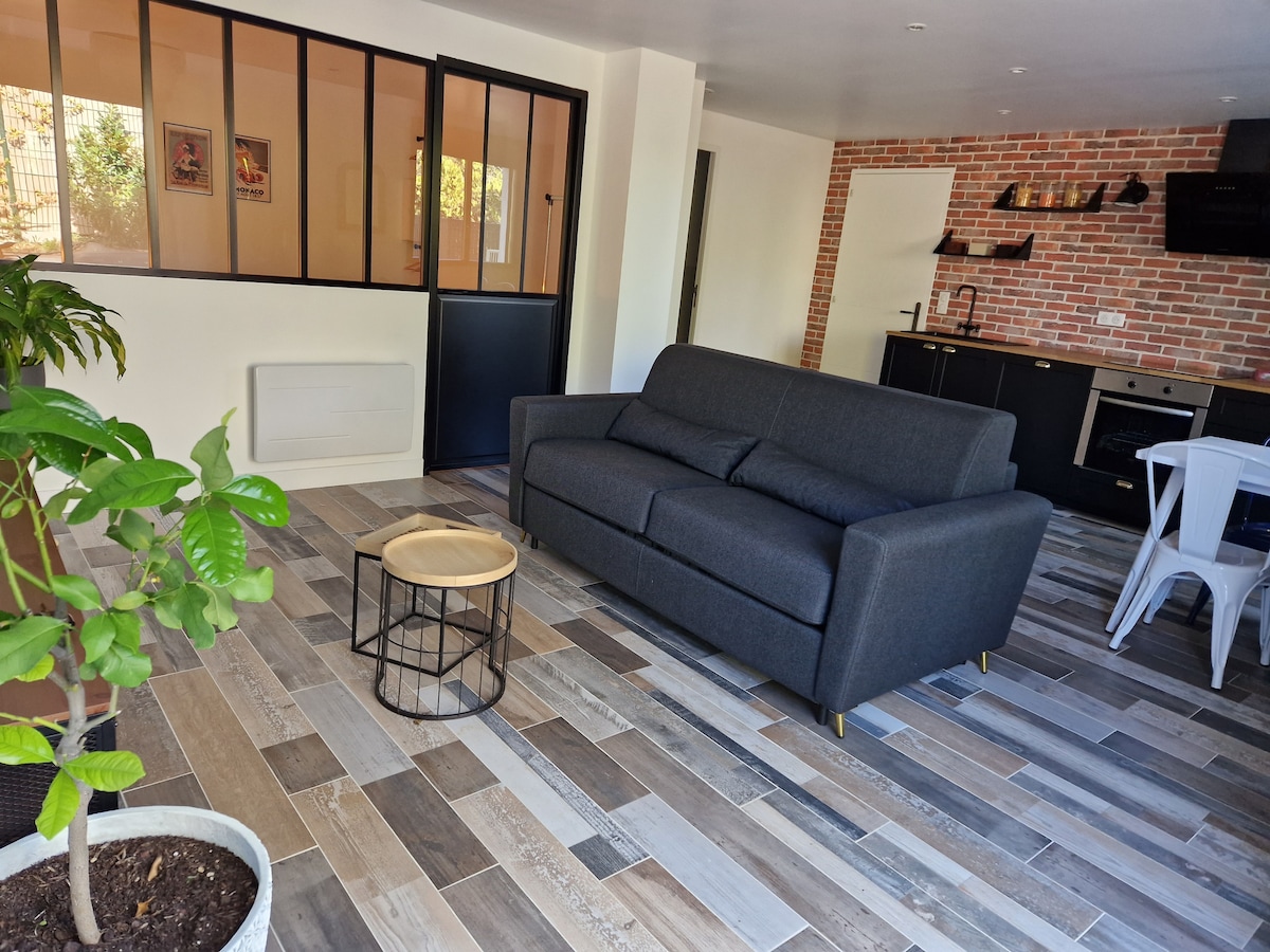 A modern living space features a gray sofa, complemented by a small round table with a black frame. The floor is covered in a variety of wooden tiles. A plant sits in a pot, and the kitchen area is visible in the background.