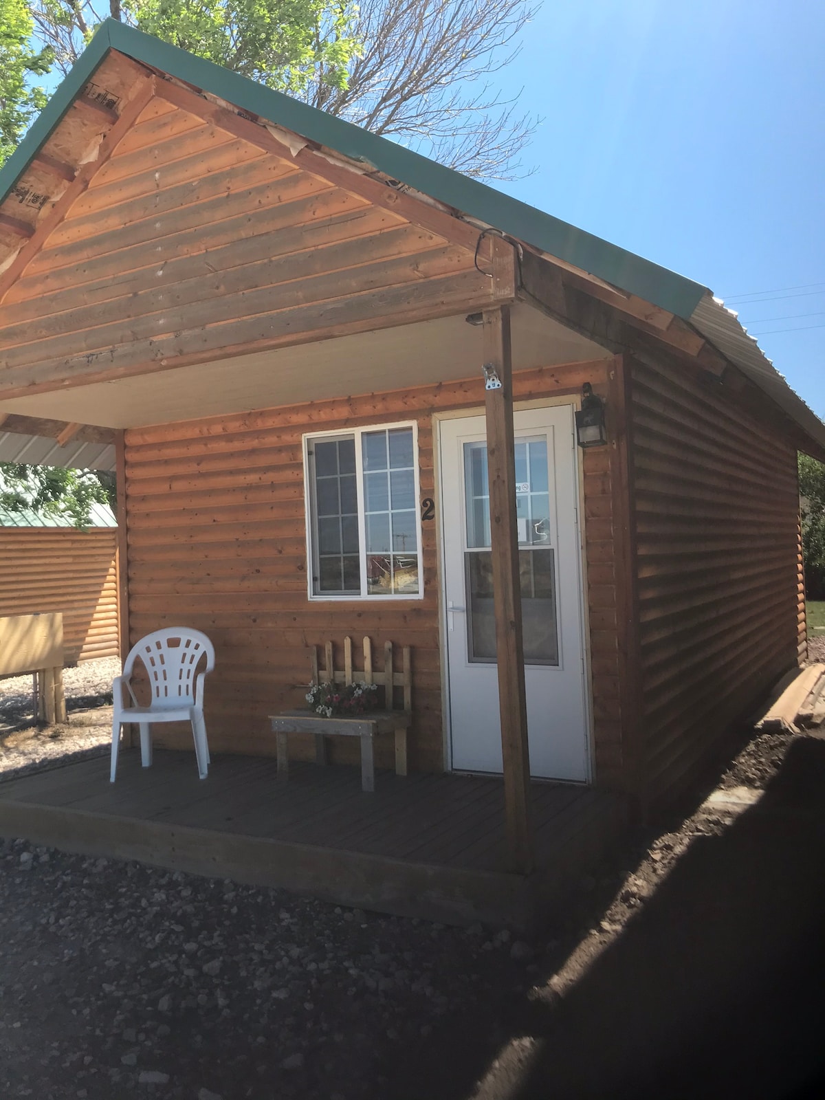 A cozy cabin exterior features a wooden facade with a sloping roof. A small porch includes a chair and a decorative bench with flowers. A window allows natural light to enter, while the surrounding area showcases gravel and additional cabins.