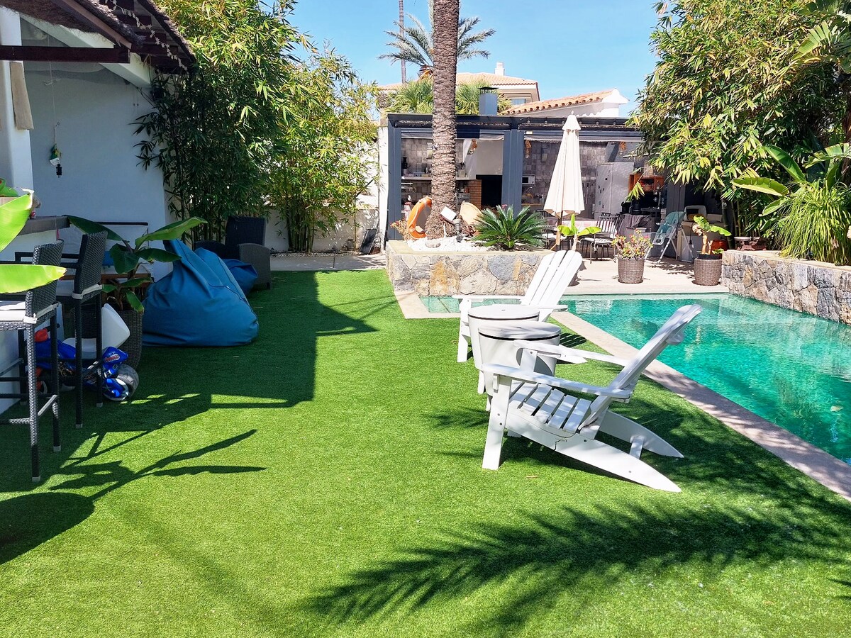 A spacious outdoor area features lush, green artificial grass, complemented by a private saltwater pool. Two white lounge chairs provide a relaxing spot, while a dining table with chairs is positioned nearby. Palm trees and plants add a tropical touch to the setting.