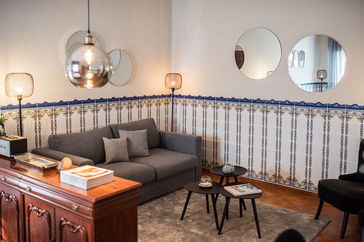 A comfortable living area is displayed, featuring a grey sofa with several cushions. Three round mirrors are mounted on the wall, reflecting soft light from stylish lamps. Decorative tiles adorn the lower part of the wall, adding a unique touch to the ambiance.