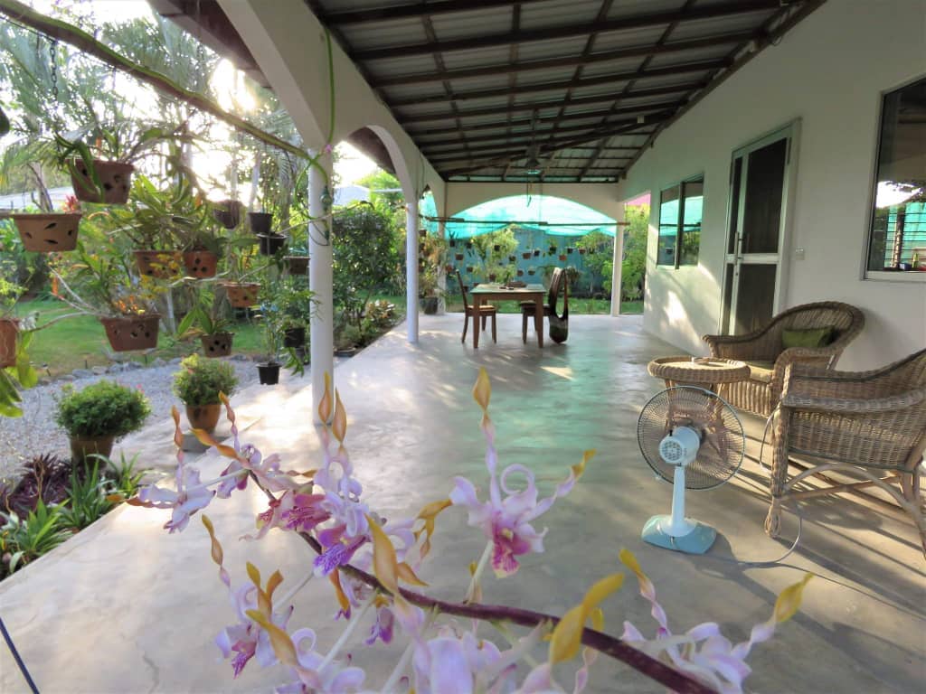 A spacious outdoor terrace is featured, surrounded by greenery and potted plants. A dining table is present, complemented by comfortable seating. A fan is visible to provide cooling, while natural light filters through the area, creating an inviting space for relaxation.