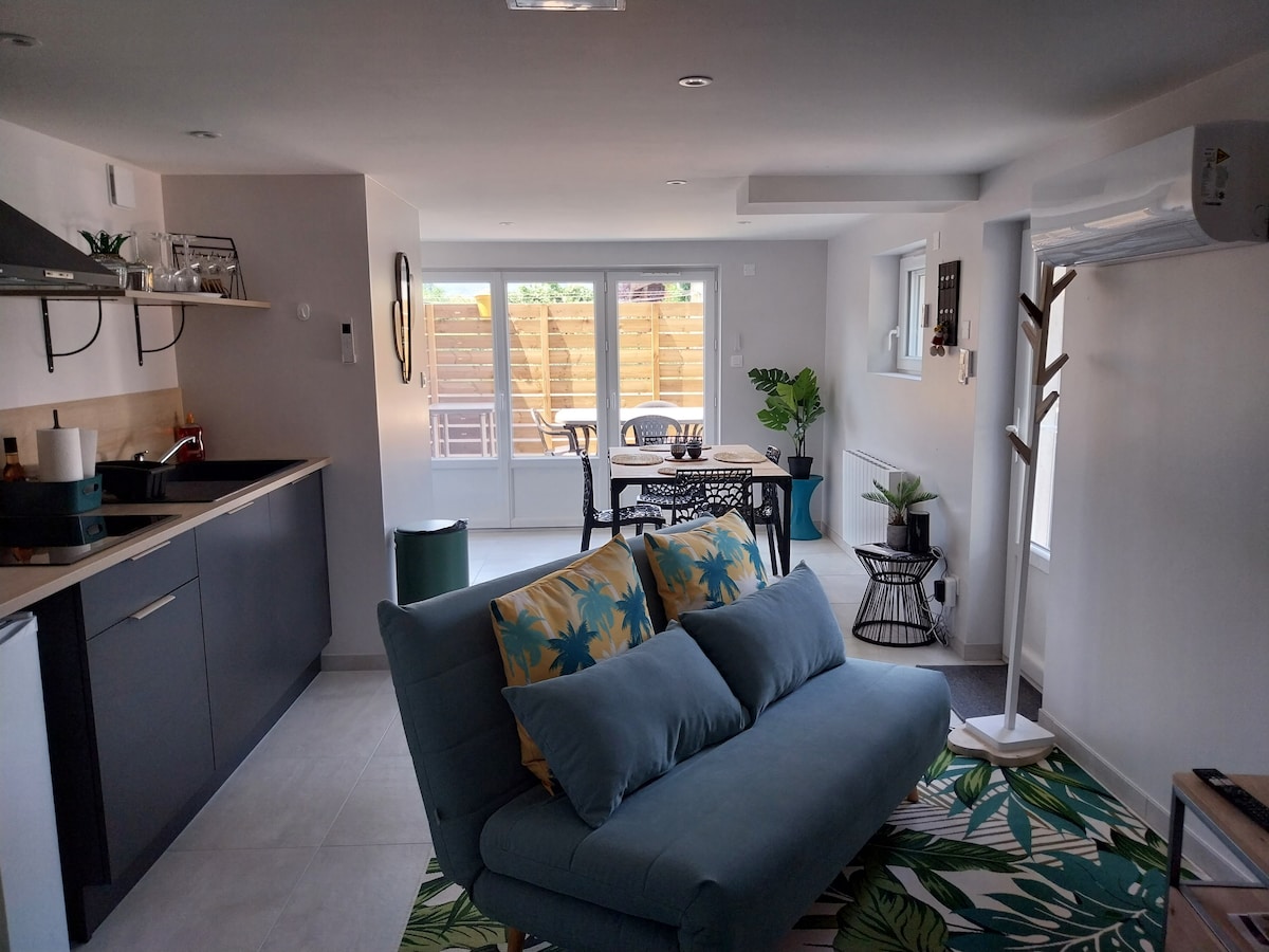 An open-plan living area features a modern kitchen with sleek cabinetry and a dining table surrounded by chairs. A comfortable sofa with colorful cushions is positioned on a patterned rug. Large windows provide a view of a private outdoor space, allowing for natural light.