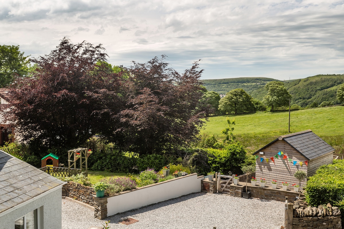 A tranquil outdoor space is depicted with a gravel area surrounded by greenery. A wooden playhouse sits to the right, accompanied by a colorful play structure. Lush trees provide shade, with expansive views of the surrounding countryside in the background.