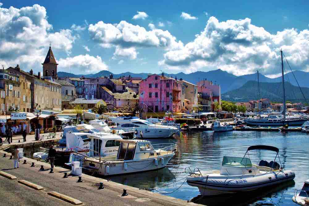 A vibrant marina scene is depicted, featuring various boats moored along the water's edge. Colorful buildings line the waterfront, with a distant mountain range visible under a cloudy sky. The atmosphere suggests a lively, coastal environment inviting leisurely exploration.