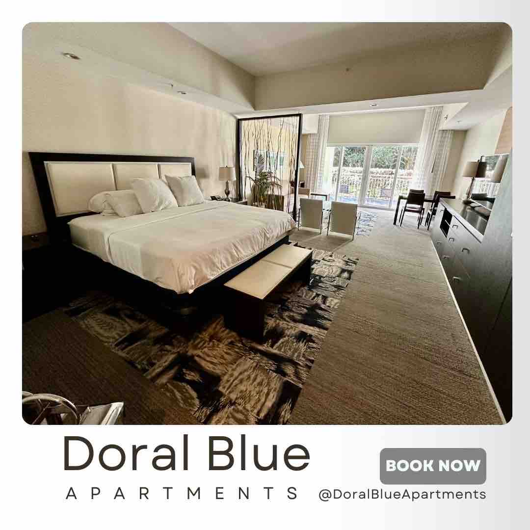 A spacious studio room features a large bed with white bedding positioned in the center. Natural light floods the area through large windows, highlighting a comfortable seating space and a sleek dresser opposite. The décor is modern, creating an inviting yet simple atmosphere.