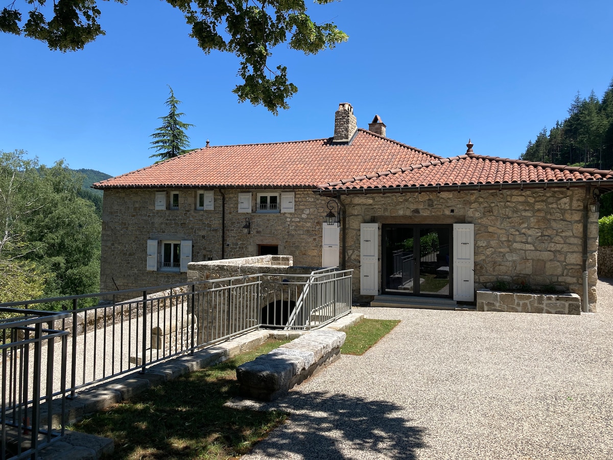 A stone house with a terracotta tiled roof is shown, surrounded by greenery and set against a clear blue sky. The building features large glass doors that open to the outdoor space, complemented by several small windows and a stone perimeter wall.