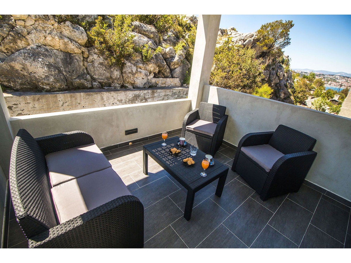 An outdoor terrace features a table with drinks and snacks, surrounded by cozy seating arrangements. The space is set against a backdrop of natural rocks and greenery, offering a serene environment for relaxation.