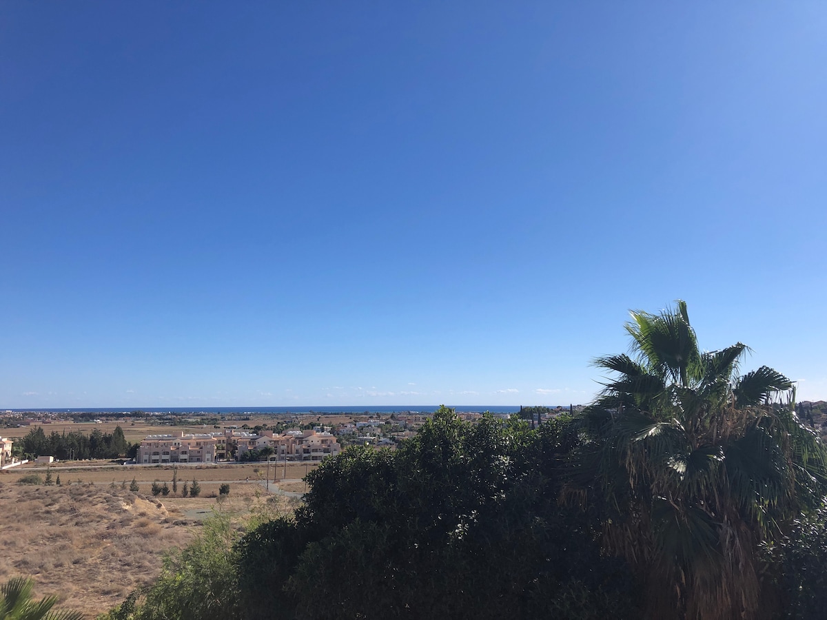 A view of the expansive landscape features clear blue skies and glimpses of the Mediterranean Sea in the distance. Lush greenery is present in the foreground, adding to the natural setting. The surrounding area includes flat terrain and scattered residential buildings.