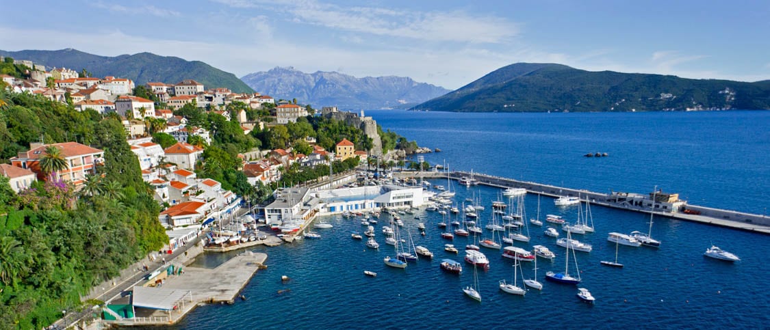 A broad view of a coastal town is shown, with white buildings and red rooftops nestled against green hillsides. A marina filled with sailboats is visible in the foreground, while the blue water extends toward distant mountains under a clear sky.