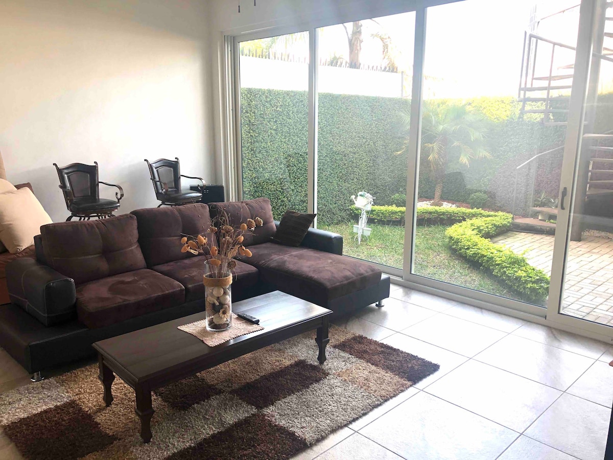 A spacious living area is observed with large glass doors that provide a view of a private garden. A dark brown sectional sofa is positioned beside a wooden coffee table, which is adorned with a decorative vase. The space is brightened by natural light and accented by a soft area rug.