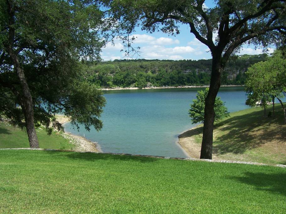 A serene view of a tranquil lake is framed by lush green grass and trees. The water reflects the blue sky and surrounding hills, creating a peaceful outdoor scene. Gentle slopes lead to the water's edge, enhancing the natural beauty of the landscape.